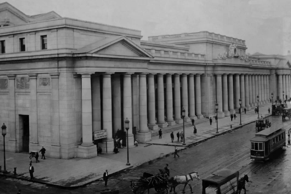 An image of historic Penn Station when it was first built in 1910, viewed from the outside with people, carriages, and streetcars passing by.